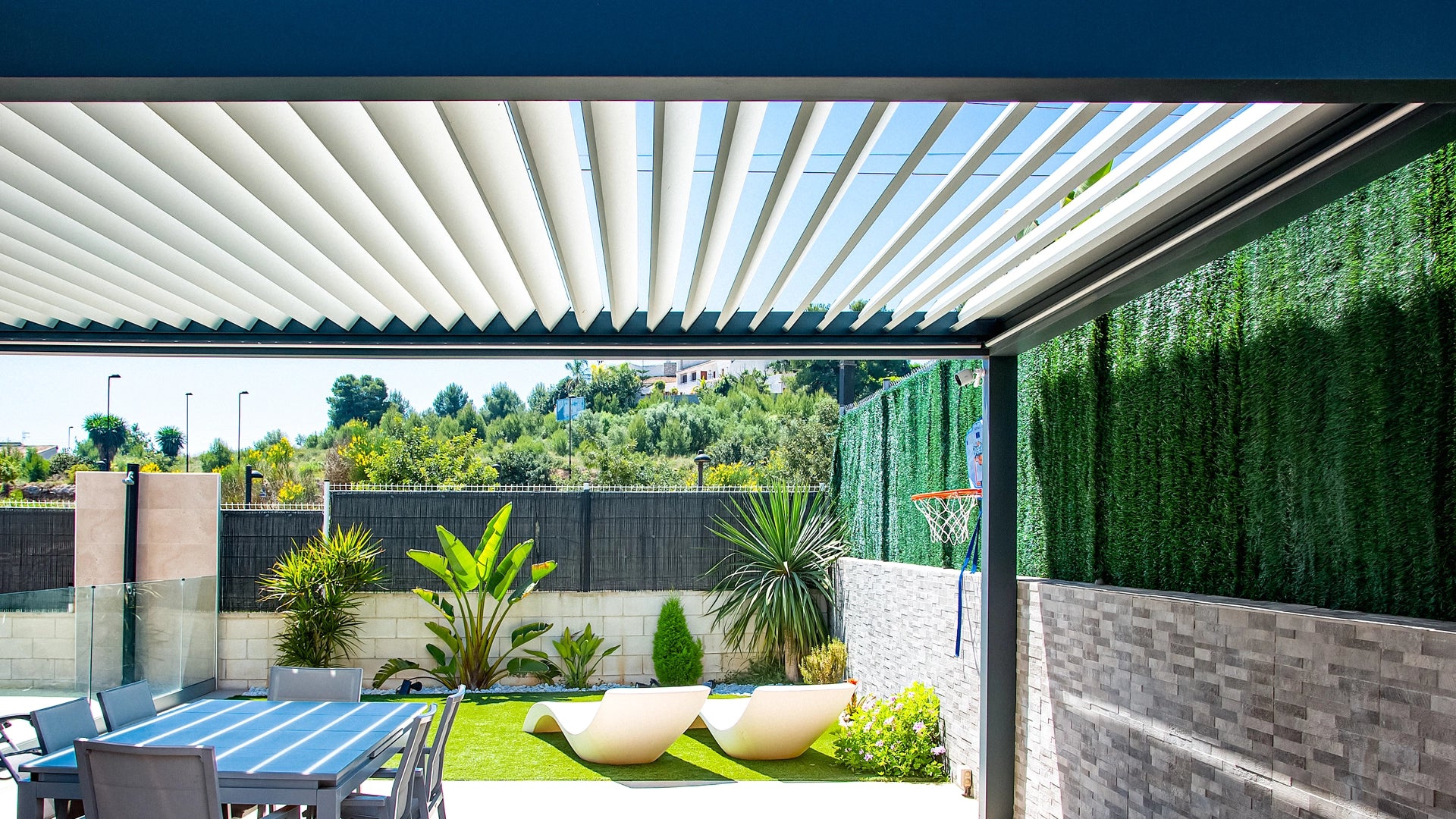 Pérgola bioclimática para jardín, terraza con mesas y sillas que da al cesped con dos tumbonas y plantas