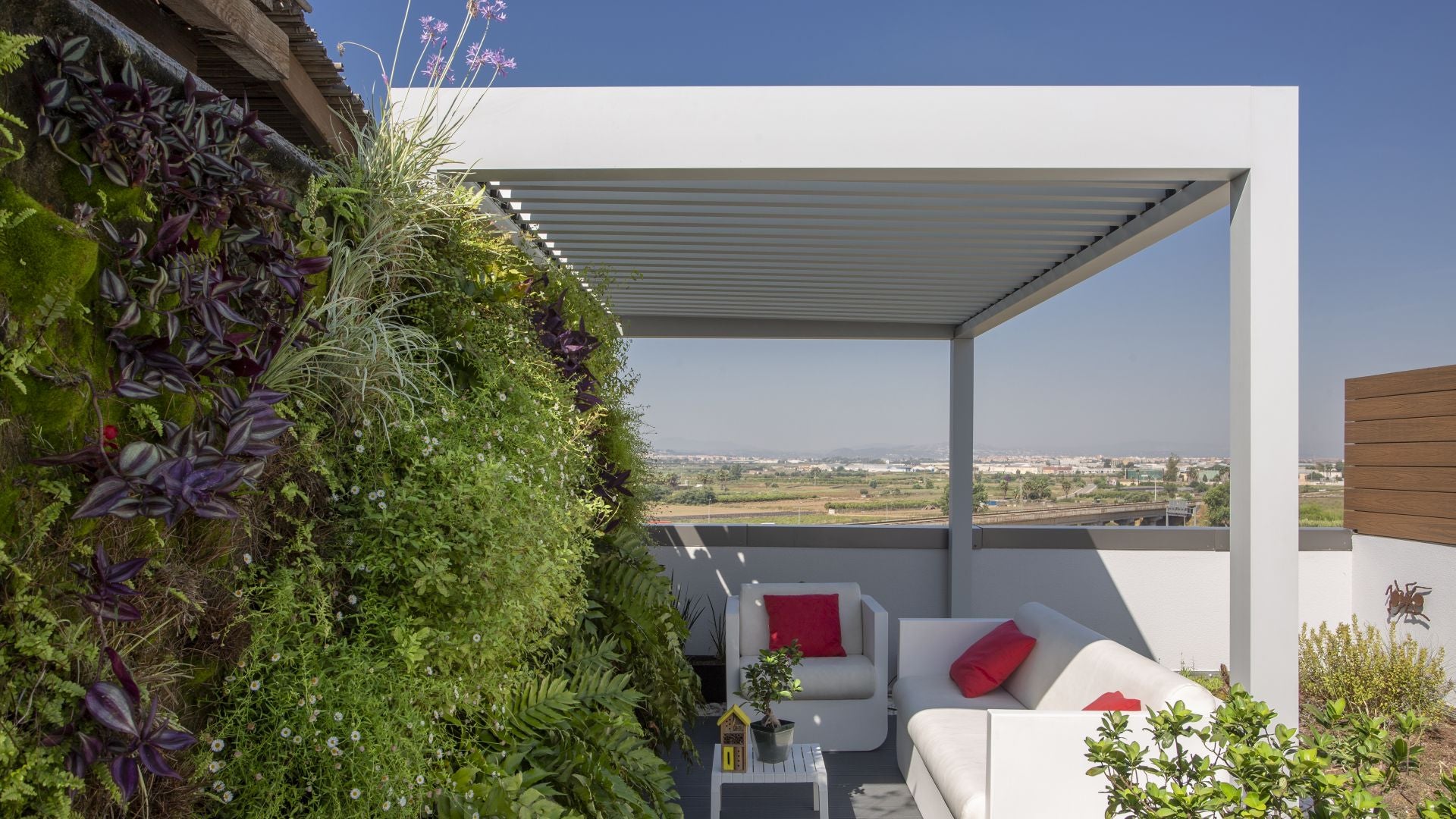 Pérgola bioclimática blanca sobre ático, con zona ajardinada y sofás y sillones blancos con cojines rojos.