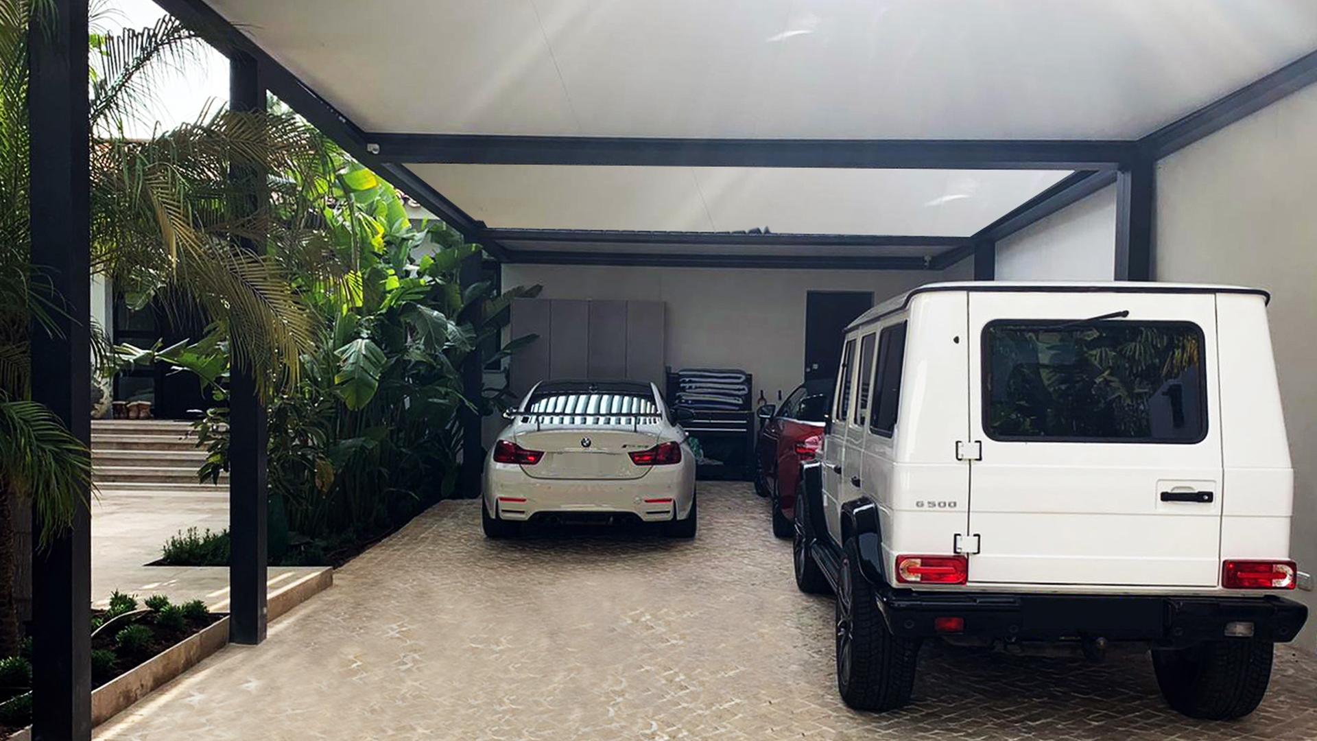 dos coches aparcados cubiertos en la parte superior y lateral por una pérgola en la entrada de una vivienda con terraza exterior.